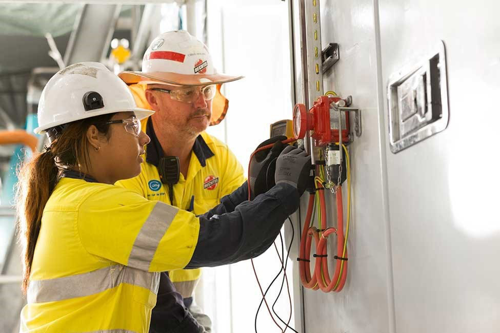An apprentice and supervisor wearing protective equipment while working together at an electrical panel